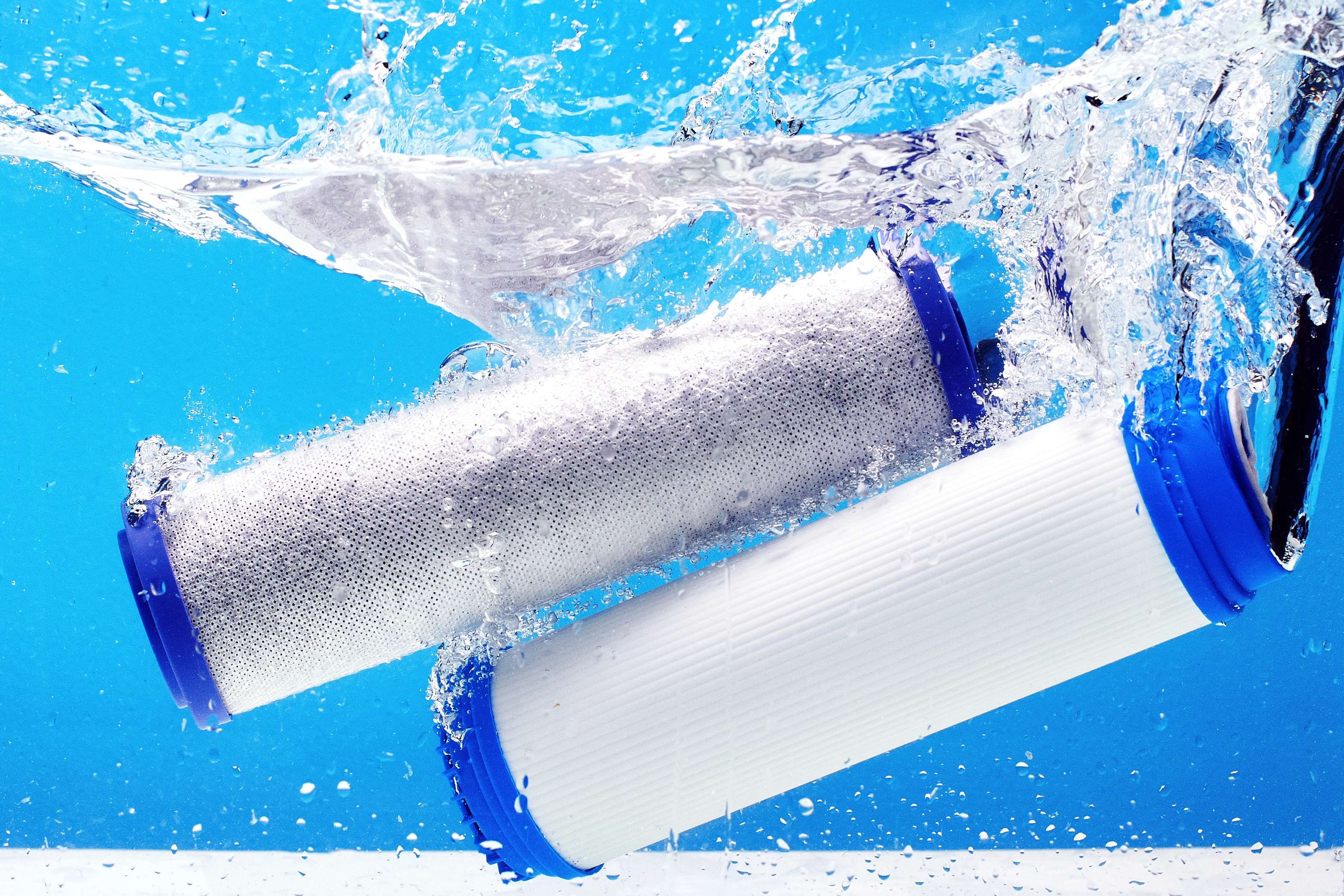 Two white cylindrical water filter cartridges with blue end caps splashing through clear blue water, surrounded by bubbles.