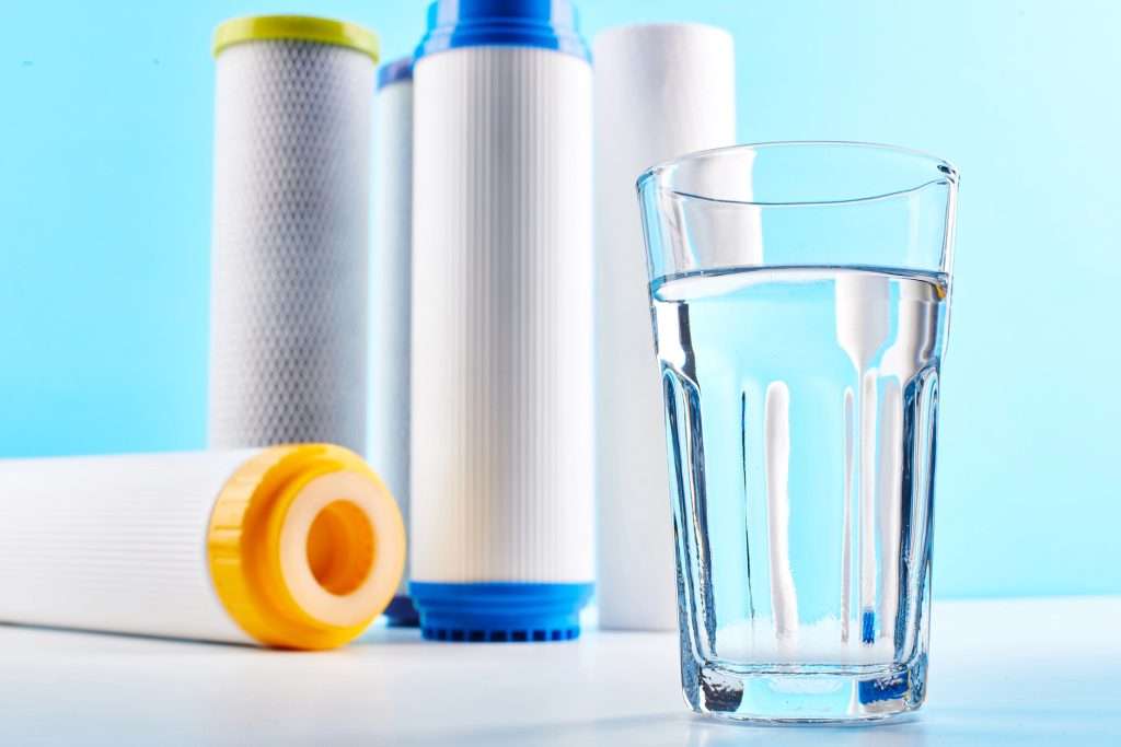 A clear glass of water resting on a white surface with five water filters in the background.