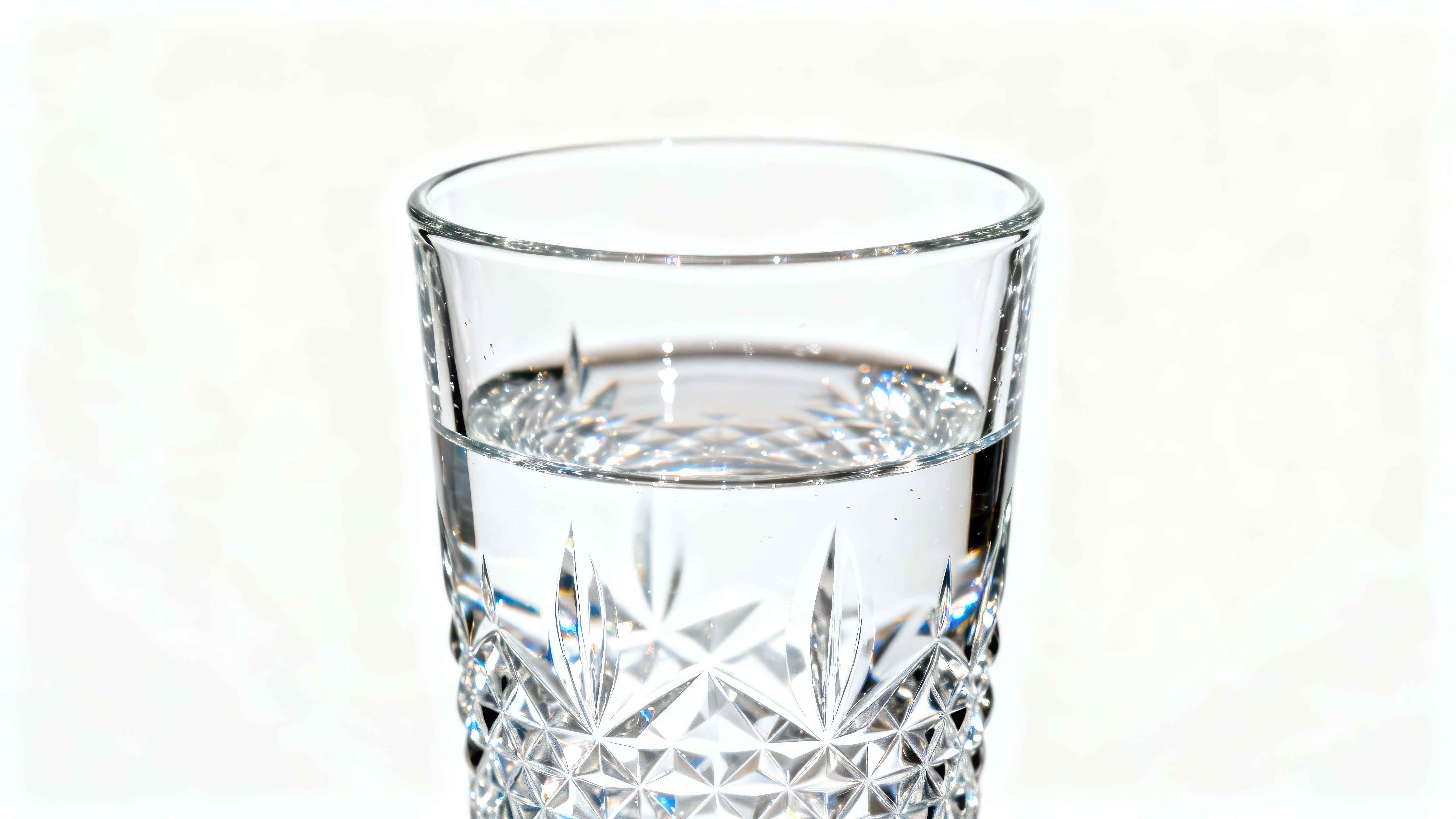 View of a drinking glass featuring intricate, diamond-cut patterns on its exterior, containing a small measure of clean, clear water.