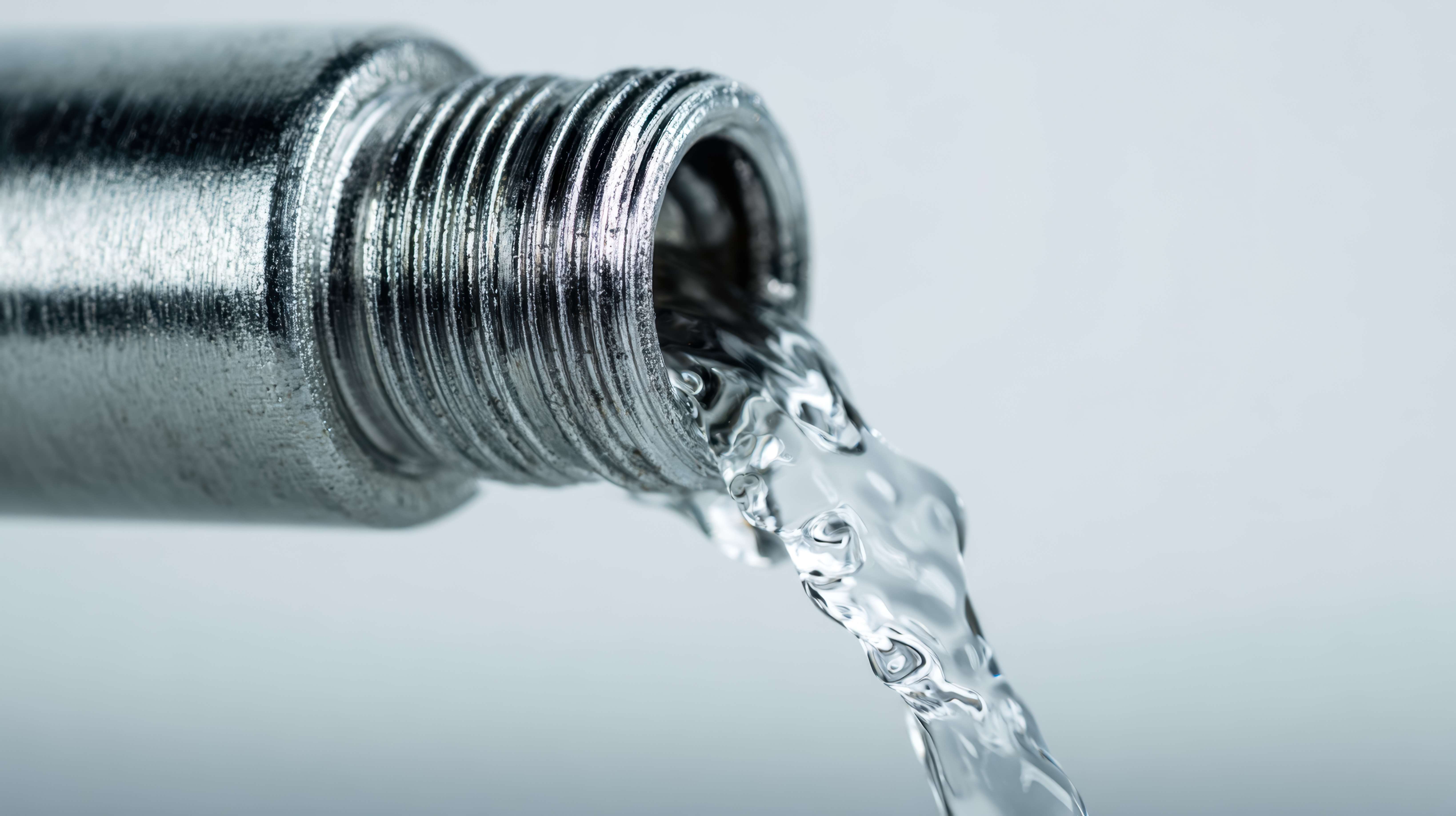 Bright, transparent water streams smoothly from a polished metal pipe with elegant spiral grooves, set against a gentle, luminous backdrop perfect for fluid studies.